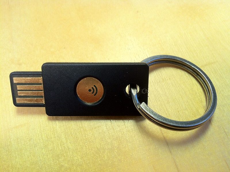 Black USB hardware security key with a round gold touch sensor, attached to a metal key ring on a wooden table