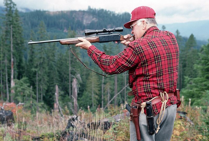 Hunter in a red plaid shirt aiming a scoped rifle in a clearing in Mt. Baker-Snoqualmie National Forest