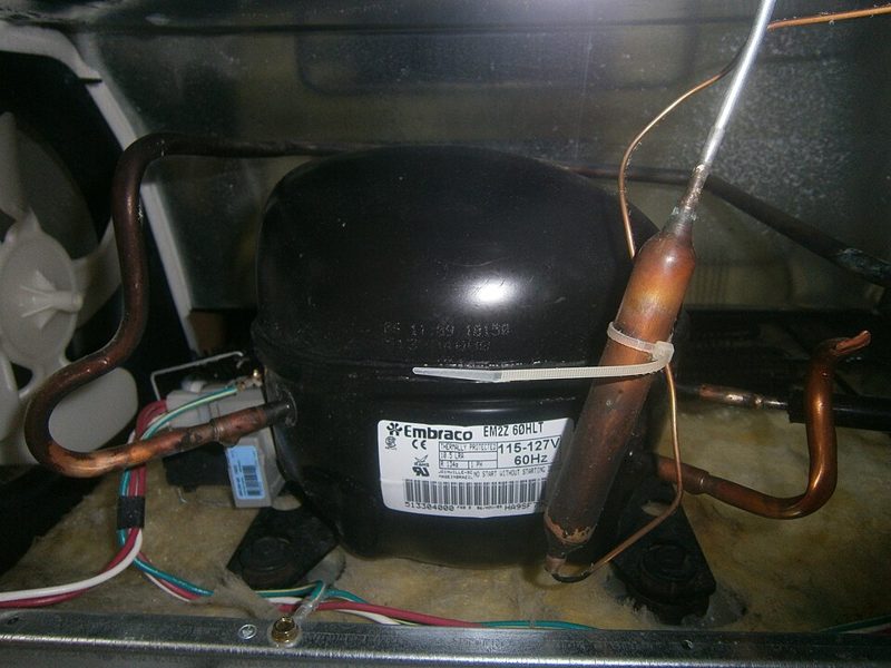 Black Embraco compressor with copper refrigerant lines at the back of a household refrigerator