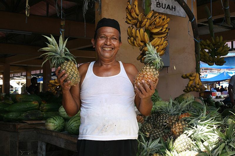 A smiling fruit seller holding pineapples at his market stall in Indonesia