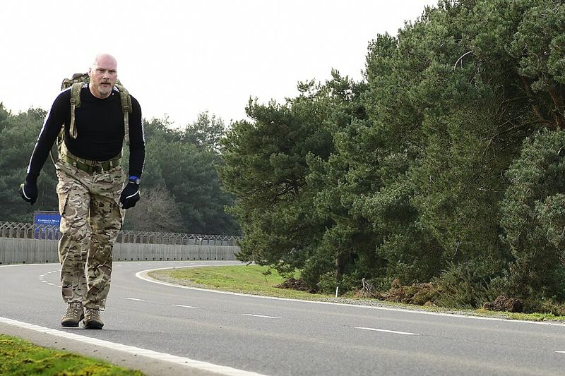 A man in camouflage trousers walking along a road while carrying a loaded military-style rucksack