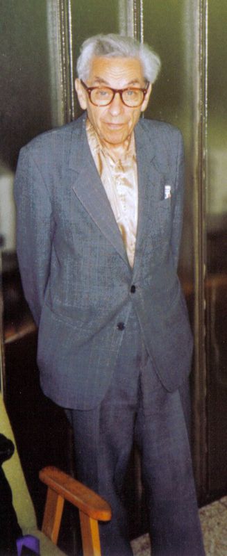 Color photo of mathematician Paul Erdős in a gray suit and glasses, standing indoors in Budapest in 1992