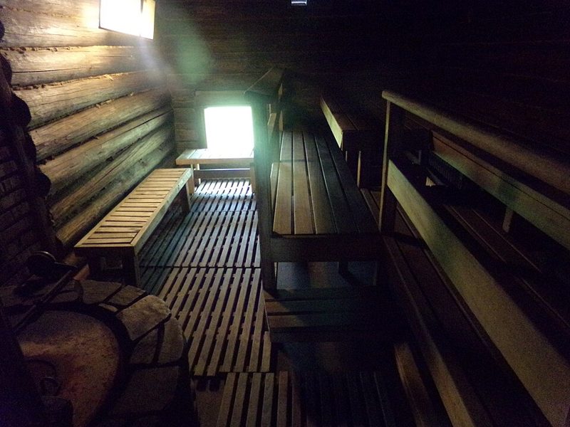 Wooden benches and slatted floor inside a traditional log sauna at Tamminiemi, Helsinki, with a stone stove in the corner