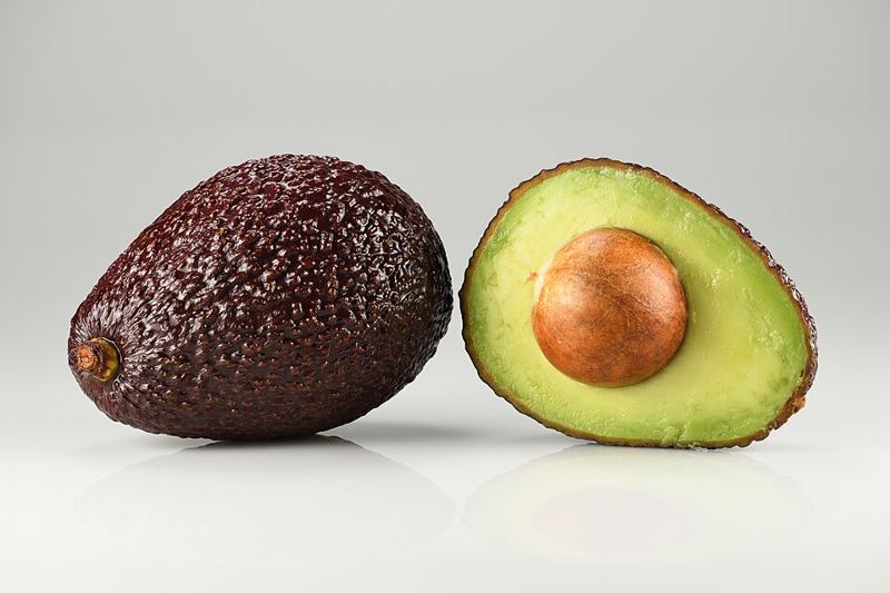 A whole Hass avocado next to an avocado cut in half with its pit