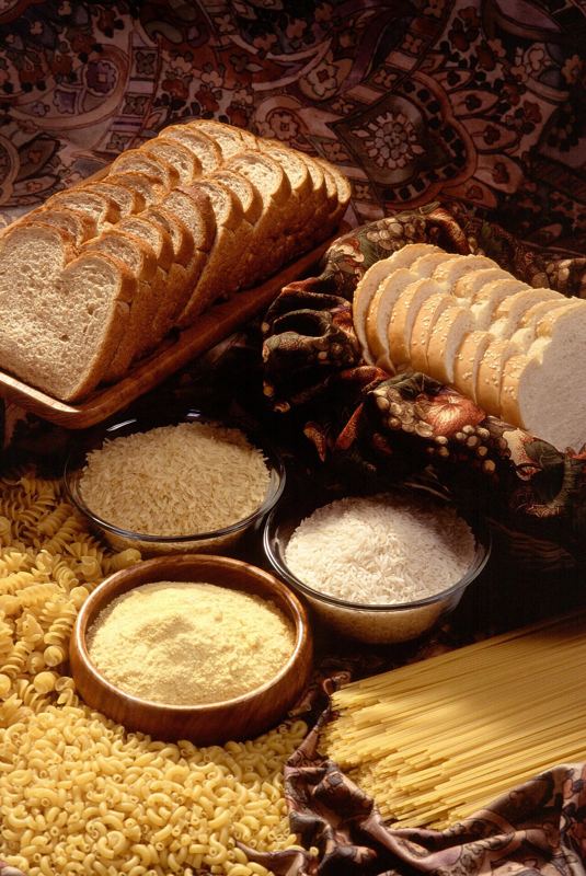 Sliced white and wheat bread, a bowl of flour, cornmeal, rice and pasta arranged on a table