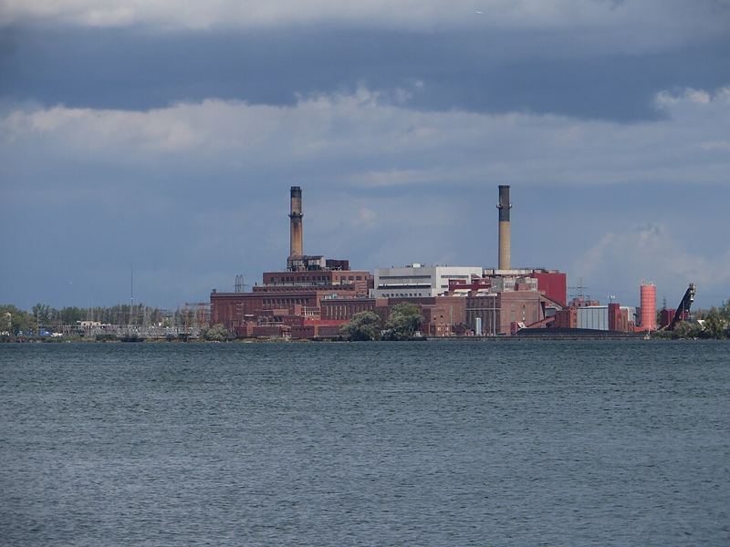 Brick power station with two tall smokestacks on a riverbank under a cloudy sky