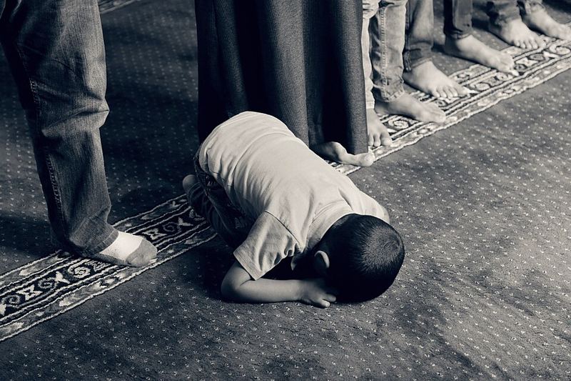 A young boy bowing in prostration during prayer on a mosque carpet beside standing worshippers