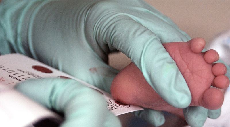A gloved health worker pricks an infant's heel to collect blood spots on a screening card for a PKU test