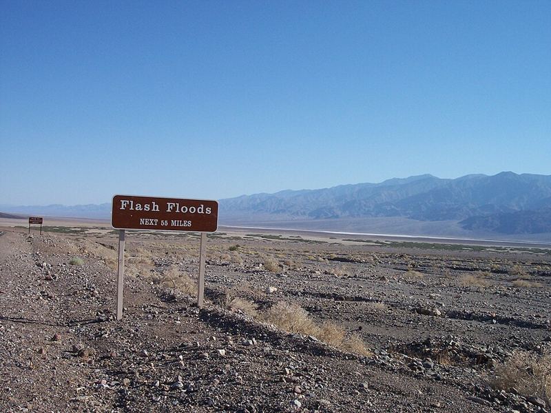 Brown roadside sign reading Flash Floods Next 55 Miles in the desert of Death Valley National Park, with mountains in the background