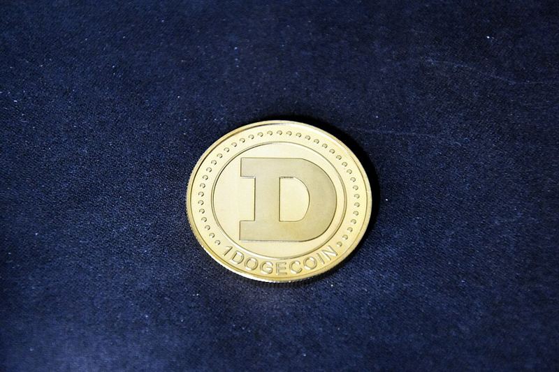 A gold-colored novelty Dogecoin coin stamped with a large D and the words 1 DOGECOIN, on a dark fabric background