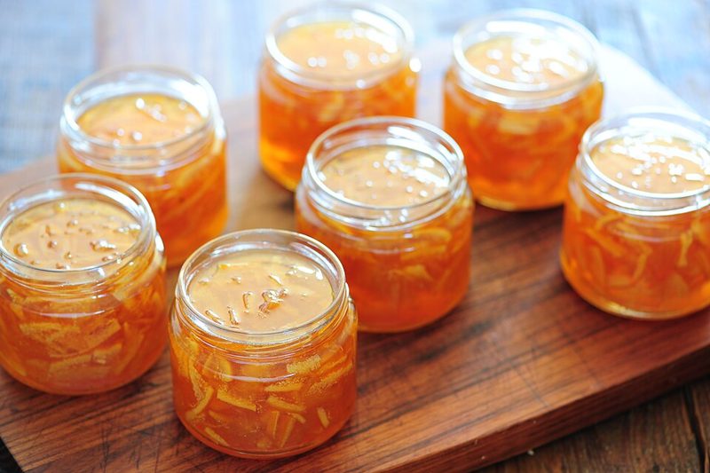 Seven small glass jars of homemade orange marmalade with visible strips of peel on a wooden board