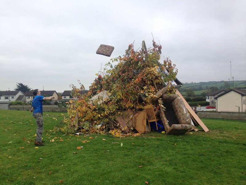 A man tossing a cushion onto a large bonfire pile of branches and old furniture on a grassy field in Ireland