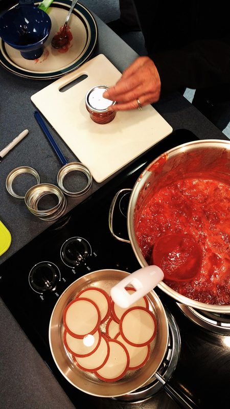A cook placing a lid on a jar of homemade strawberry jam beside a pot of cooked fruit