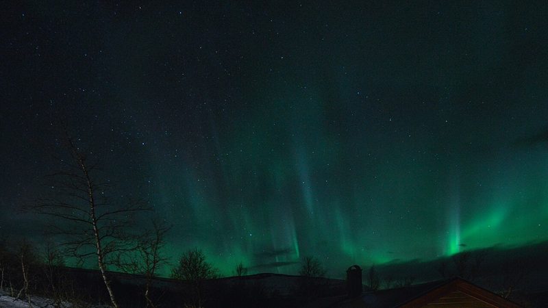 Green northern lights over a snowy hillside and cabin roof at night