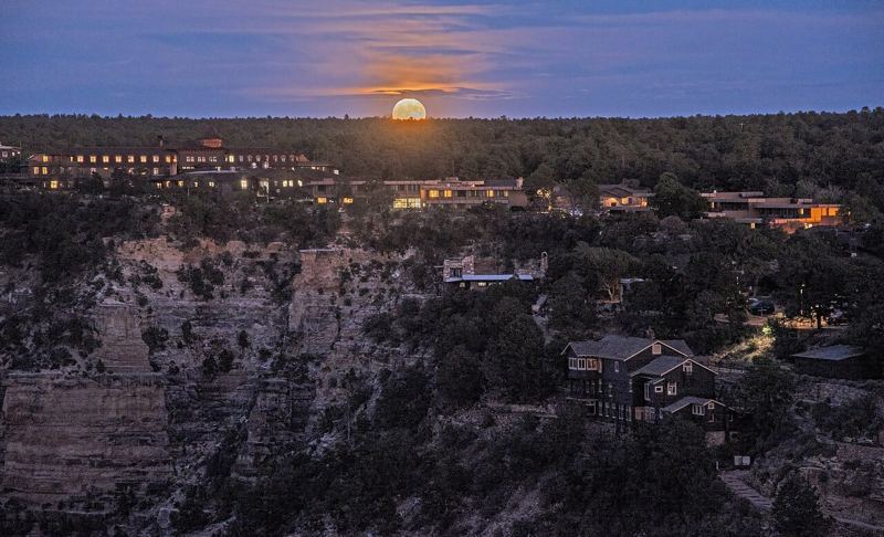 A full moon rising just above the forested horizon behind lit buildings of Grand Canyon Village at dusk